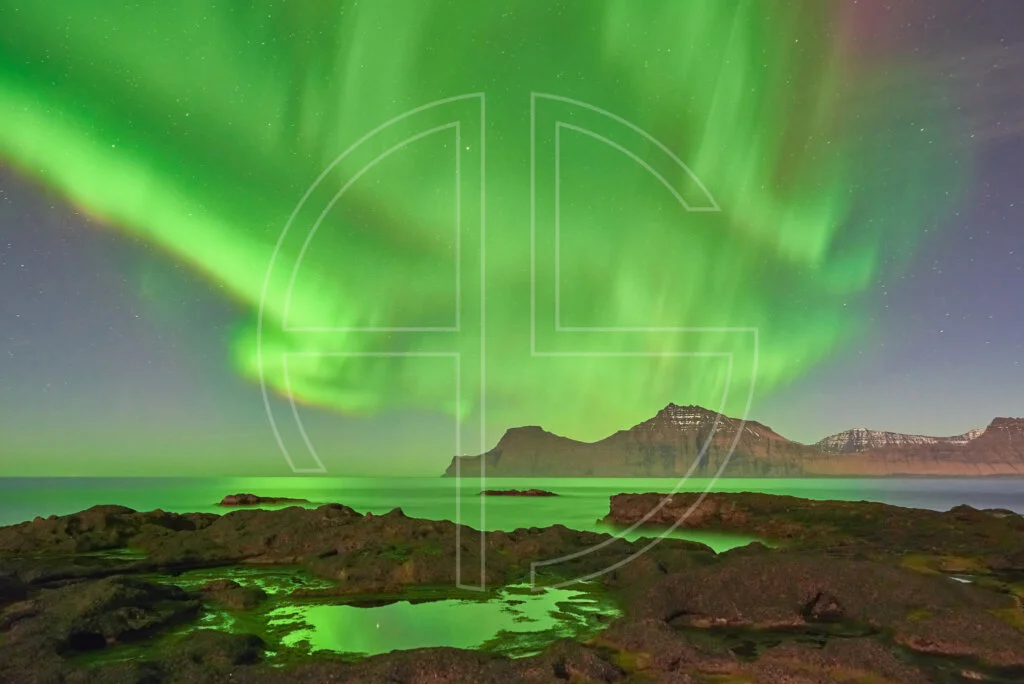 Northern Lights at the sea. The foreground has rocks with water puddles.