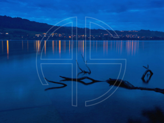 A lake at the blue hour. The lights of a town in the background reflect on the water.