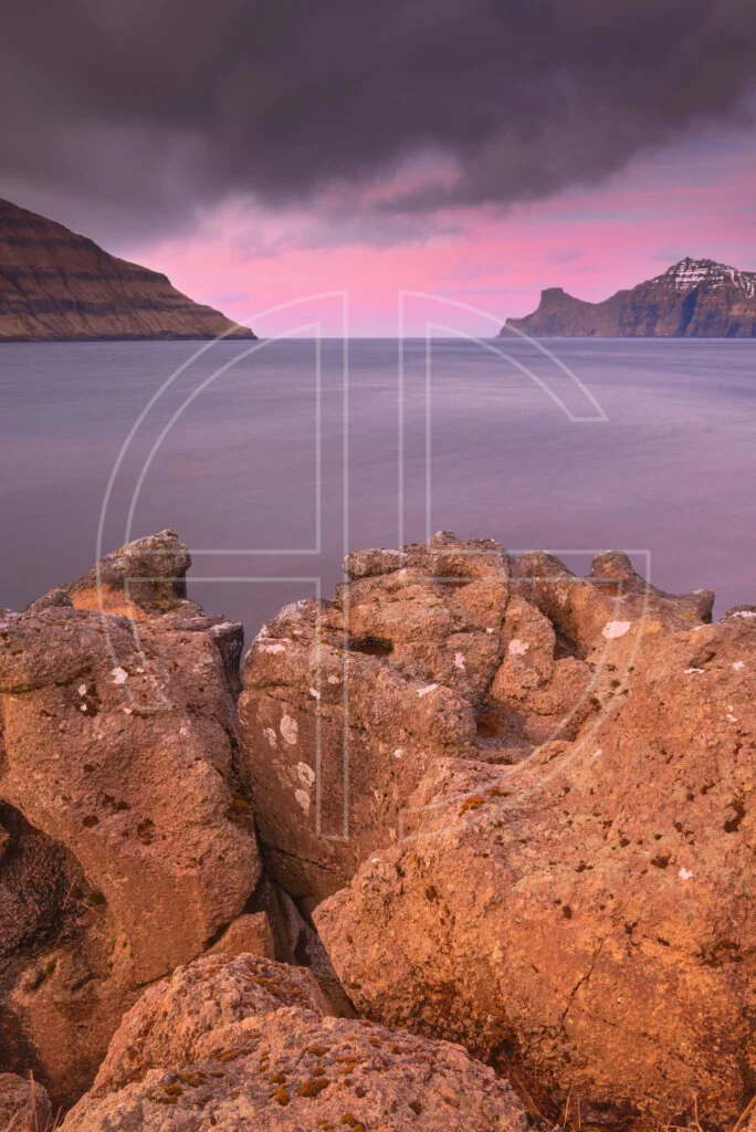 Pink dusk at the sea under a dark sky. In the foreground lie rocks.
