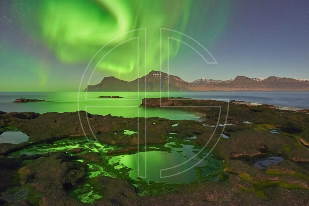 Northern Lights at the sea. The foreground has rocks with water puddles.