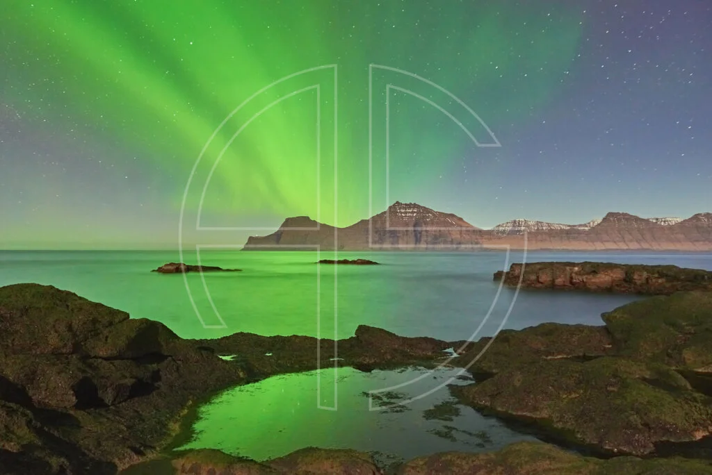 Northern Lights at the sea. The foreground has rocks with water puddles.
