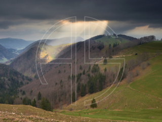 Beams of light break through black clouds, illuminating hills.