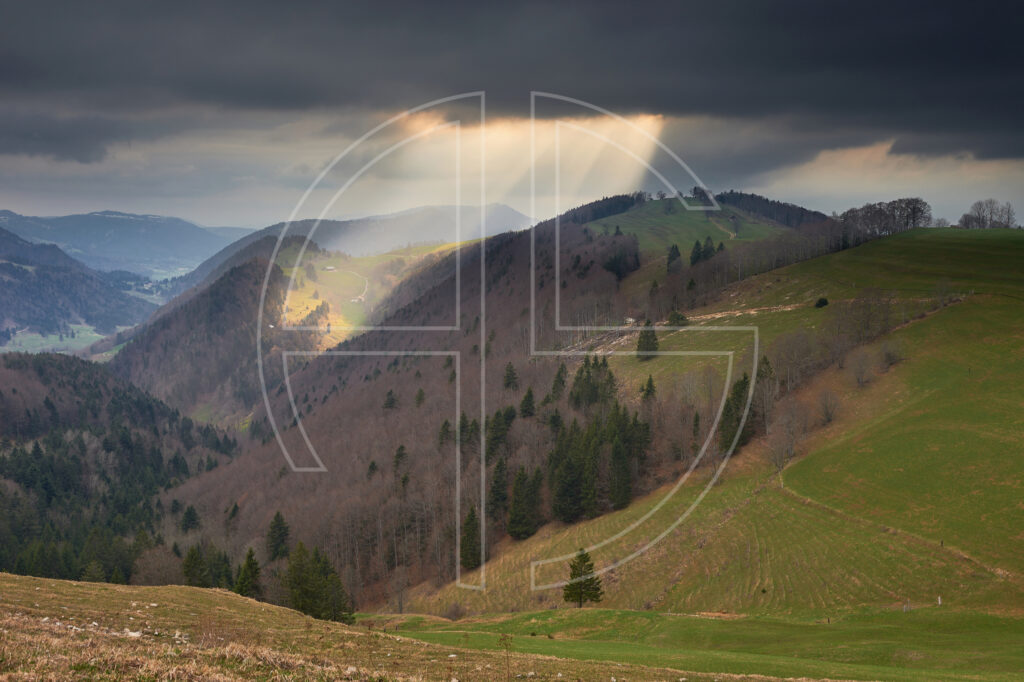 Beams of light break through black clouds, illuminating hills.